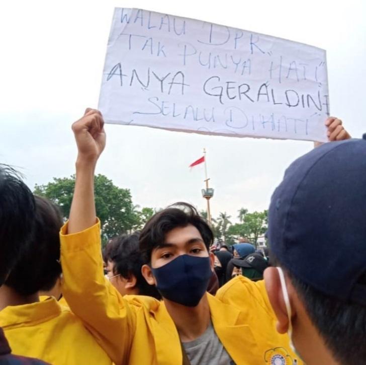 tulisan spanduk yang ditunjukkan lelaki yang mengenakan almamater warna kuning salah satu universitas.
